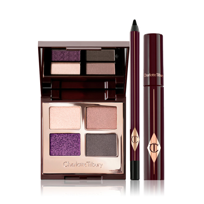 An open, mirrored-lid eyeshadow palette with eyeshadows in shades of purple, gold, and grey, an eyeliner pencil in black, and a black mascara in dark crimson packaging.