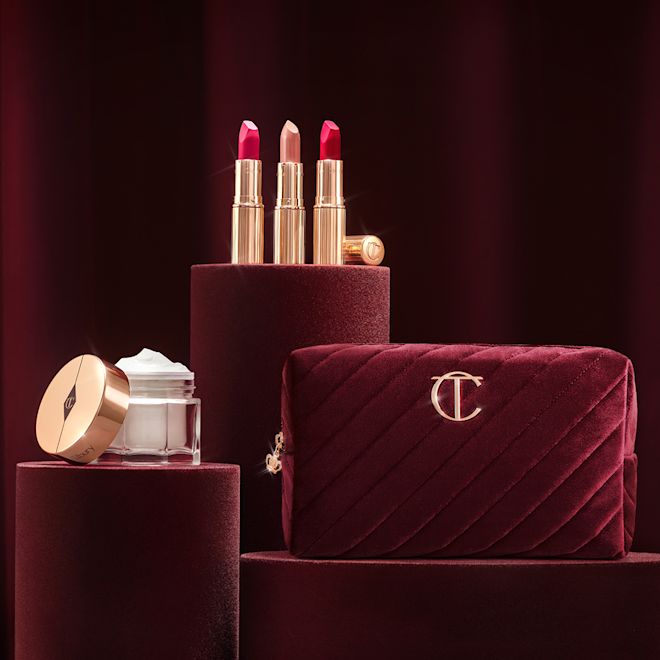 Platinum Jubilee Collection Banner with a red velvet makeup bag, pearly-white face cream in a glass jar with a gold-coloured lid, and three open lipsticks, two with a matte finish in magenta and raspberry pink and a satin finish one in a soft rosy peach shade.