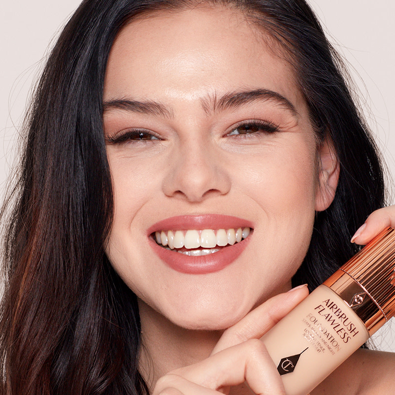 A fair-tone model with a glowy, smooth face base and wearing matte, muted red lips and nude eye makeup while holding a foundation bottle. 