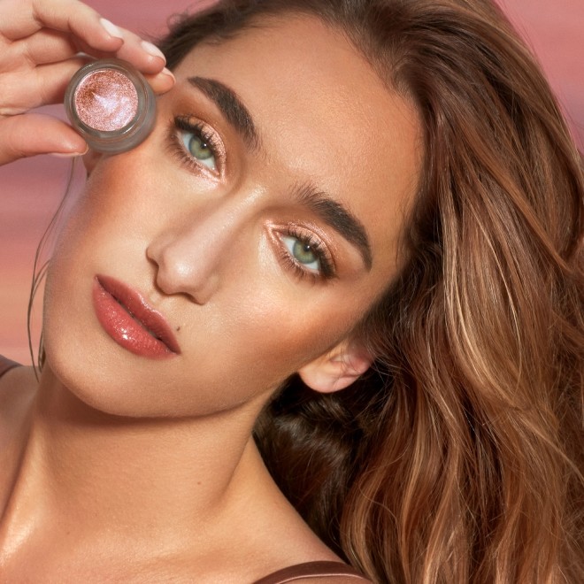 Light-tone model with blue eyes wearing golden pink duo-chrome-effect cream eyeshadow with a duo-chrome metallic finish while holding up the eyeshadow pot next to her face.