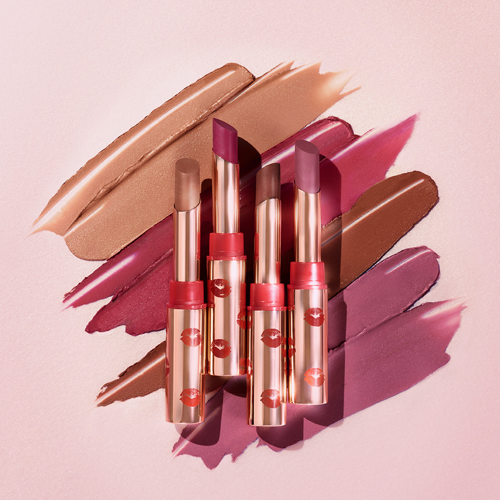 Banner with four open matte lipsticks in fuchsia, peachy-beige, dark mauve, and chocolate brown in gold-coloured tubes with red-coloured kiss print all over. 
