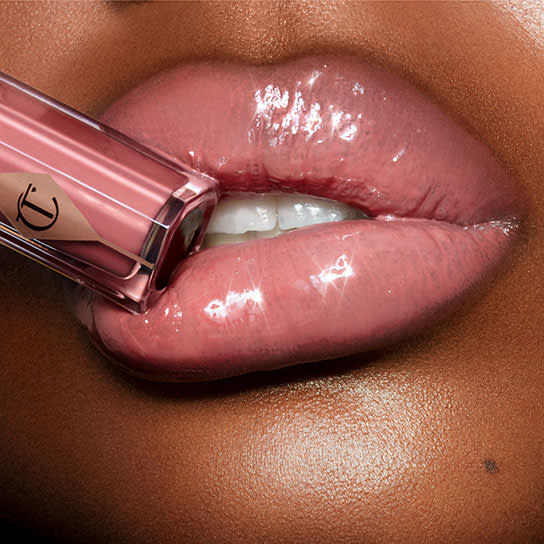 Lips close-up of a deep-tone model wearing a high-shine, nude-pink gloss and holding the lip gloss tube to her lips. 