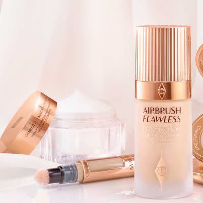 A thick white cream in an open glass jar with the lid resting on the side with an opened concealer laying flat and a bottle of beige-coloured substance with a rose-gold cap.