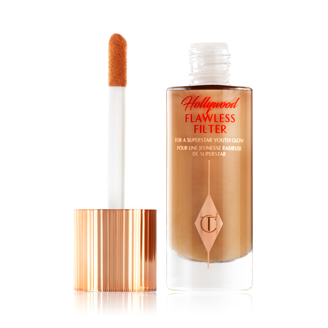 An open, luminous primer in a dark golden-beige shade in a glass bottle with its gold and white doe-foot applicator next to it.