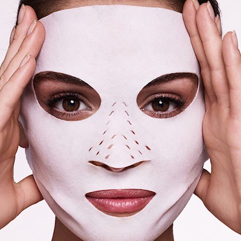 Close-up of a light-tone, brunette model wearing a sheet mask made of textile fabric with eyes, lips, and nose openings.