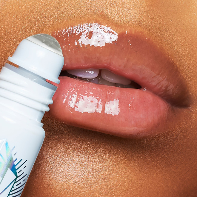 Close-up of a deep-tone model applying a nourishing, clear lip oil with a crystal roller ball applicator. 