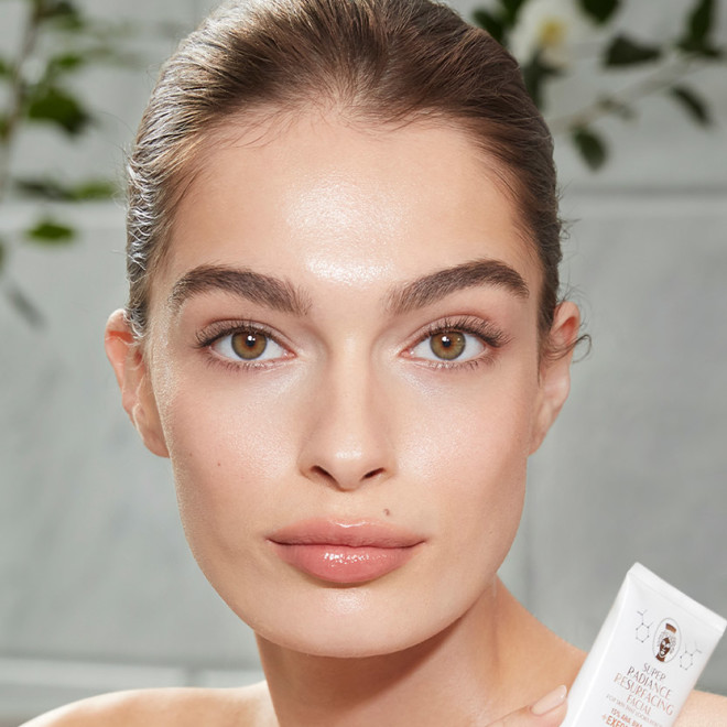 Light-tone model with hazel eyes and smooth, flawless, and glowy glass skin holding a white-coloured tube of a wash-off exfoliating mask.
