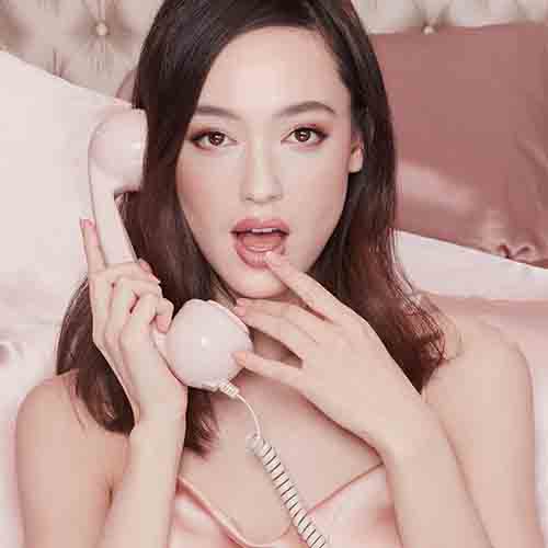 A fair-tone model wearing glowy, nude pink makeup and a nude-pink satin dress while holding a blush pink corded phone. 