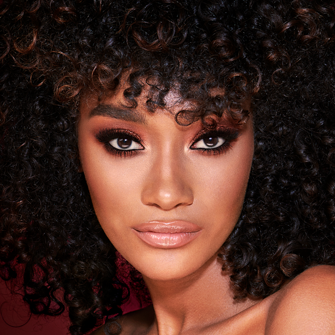 The Bella Sofia Look on deep tone model with brown eyes A deep-tone model with brown eyes wearing smokey brown and gold eye makeup with warm pink blush and glossy nude-pink lips