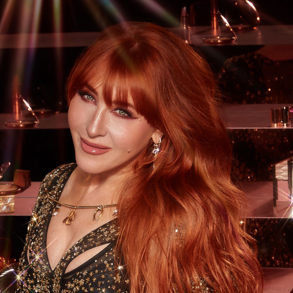Charlotte Tilbury wearing nude pink lipstick with glowy pink blush, and berry pink eye makeup with dangly earrings, delicate necklace, and sparkly dress. 