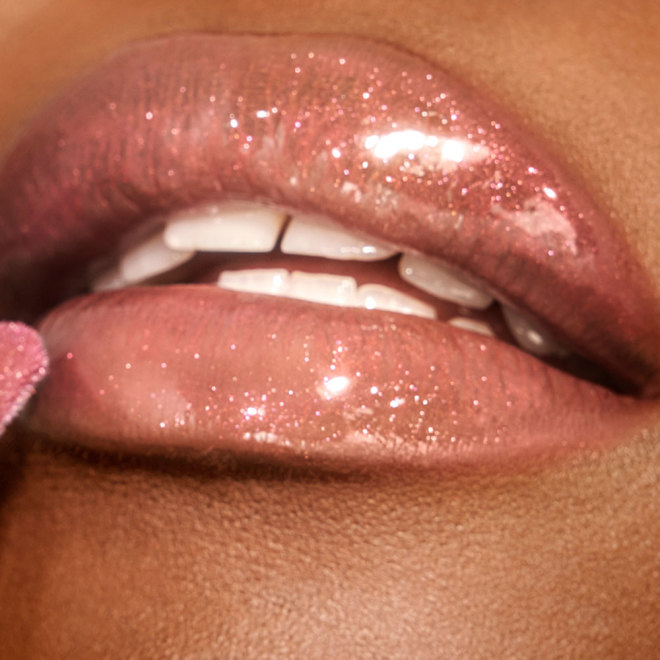 Lips close-up of a deep-tone model applying a shimmery lip gloss in a rosy opal shade with fine glitter.