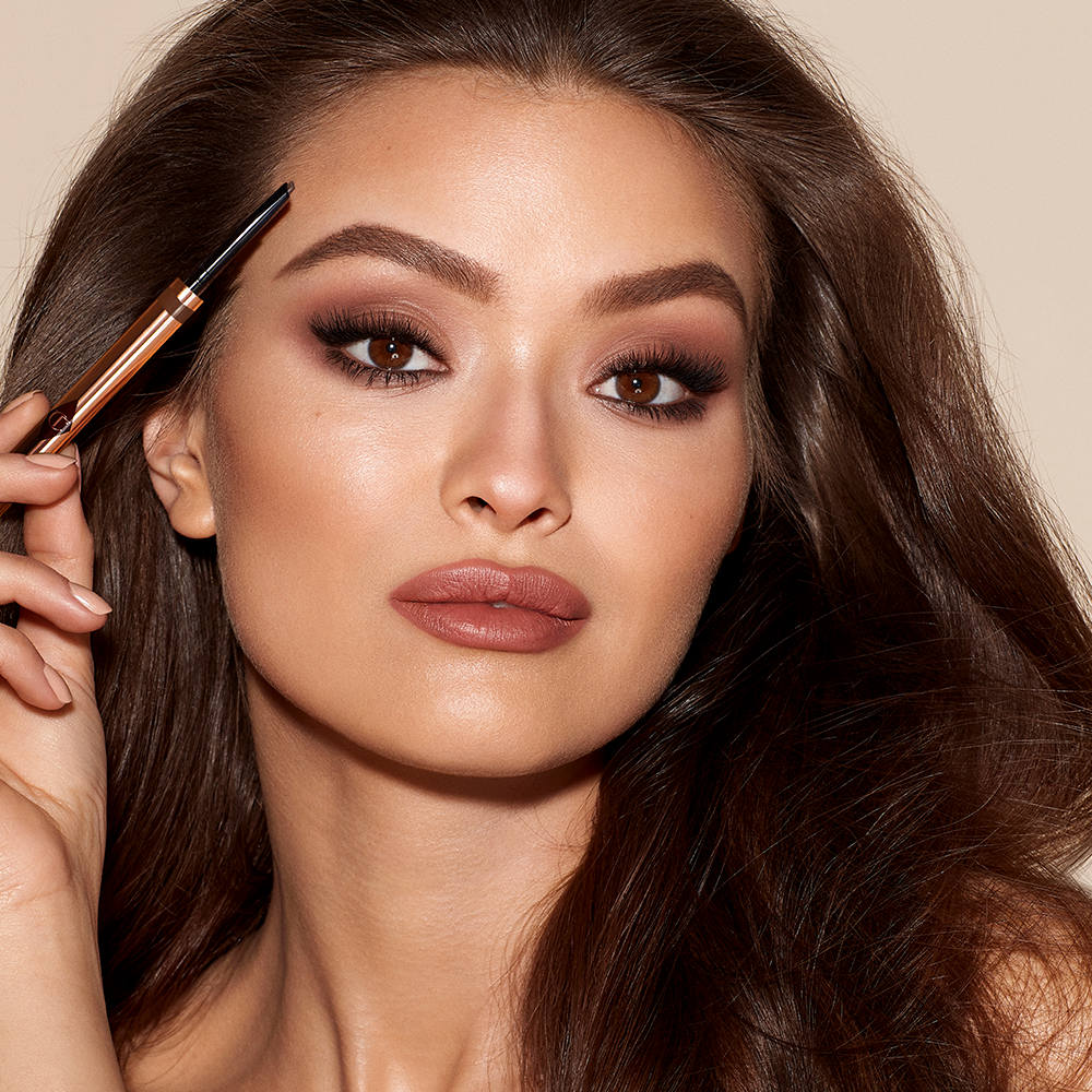 Brow Cheat in Medium Brown on medium-tone model A medium-tone model with brown eyes wearing smokey brown eye makeup with a muted brick-red lipstick and applying a medium-brown-coloured brow gel.