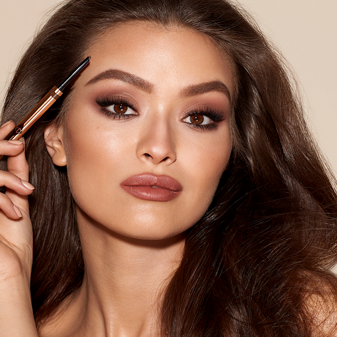 Brow Cheat in Medium Brown on medium-tone model A medium-tone model with brown eyes wearing smokey brown eye makeup with a muted brick-red lipstick and applying a medium-brown-coloured brow gel.