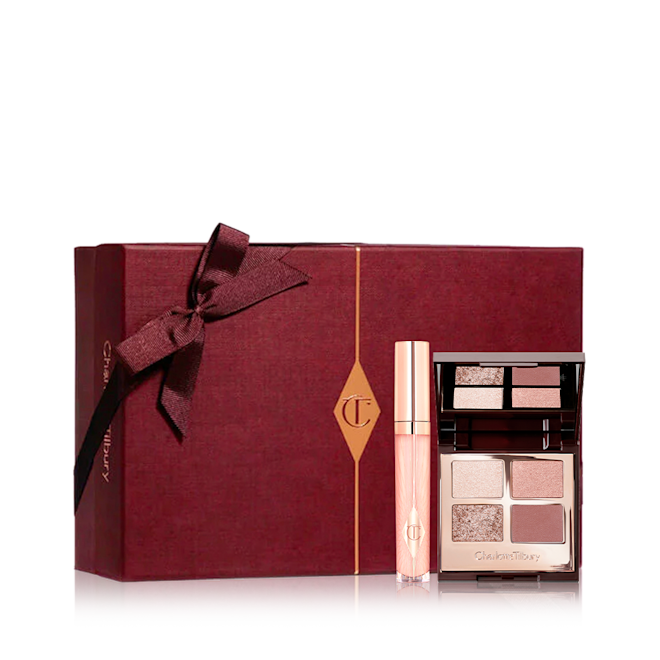 Hollywood Secrets Mystery Box displayed Sheer pink lip gloss in a glass tube with a gold-coloured lid with an open, mirrored-lid eyeshadow palette with matte and shimmery eyeshadows in brown, pink, and gold shades with a dark crimson-coloured gift box in the back.