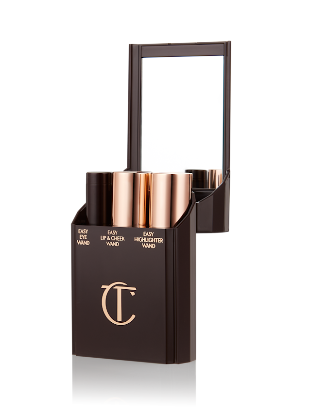 Quick & Easy Instant 5 Minute case open An open, black-brown-coloured portable makeup case with a mirrored lid, with three product places, which are filled with an eyeshadow stick, lip and cheek stick, and highlighter stick.