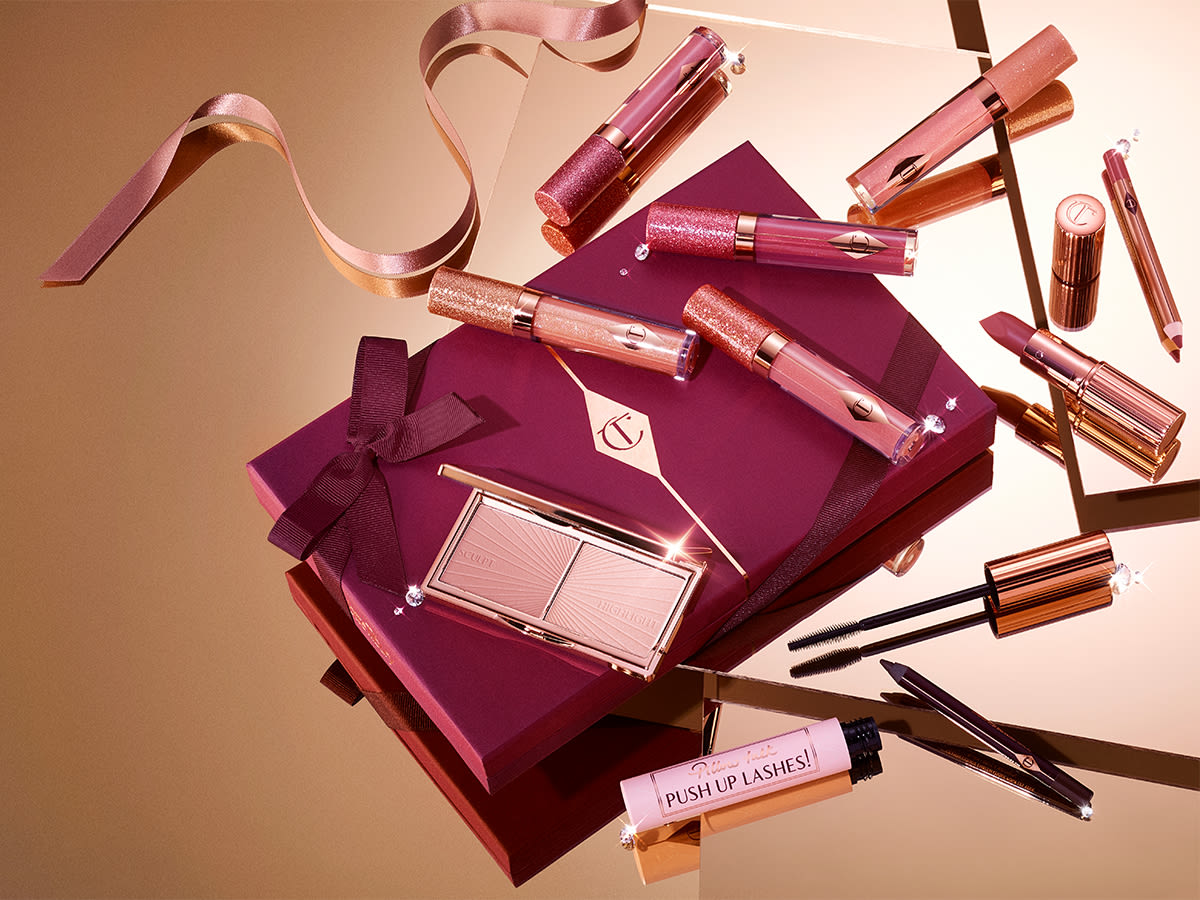 Lip, face, and eye gifts banner A collection of lip glosses, lipsticks, blush, highlighter palette, eyeliners, and mascaras on top of a dark plum gift box with ribbons scattered all around.