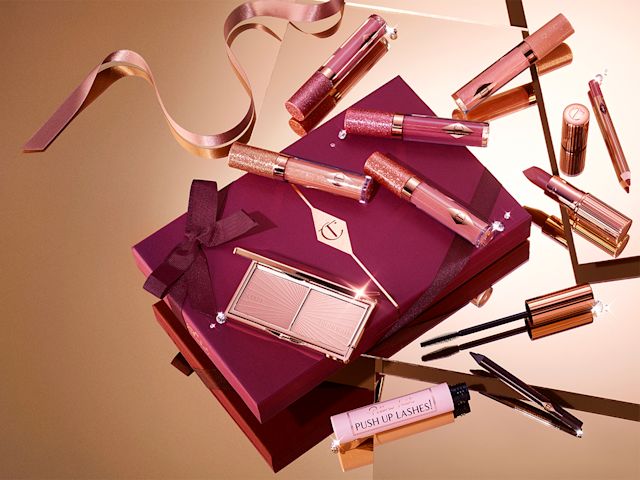 Lip, face, and eye gifts banner A collection of lip glosses, lipsticks, blush, highlighter palette, eyeliners, and mascaras on top of a dark plum gift box with ribbons scattered all around.