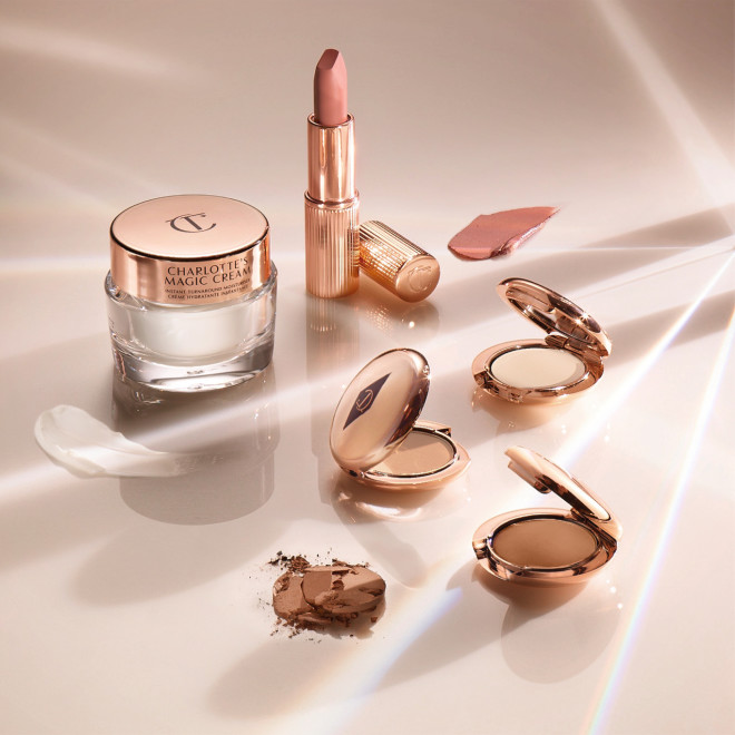 Banner with a pearly-white face cream in a glass jar with a gold-coloured lid, a collection of pressed powder compacts in fair, tan, and deep shades, and a dusky pink lipstick in a sleek gold-coloured tube.