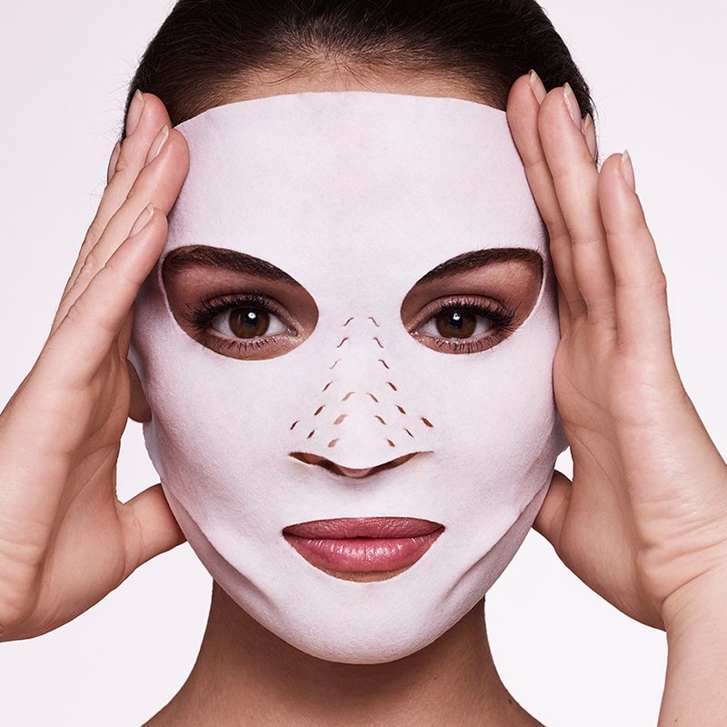 Close-up of a light-tone, brunette model wearing a sheet mask made of textile fabric with eyes, lips, and nose openings. 