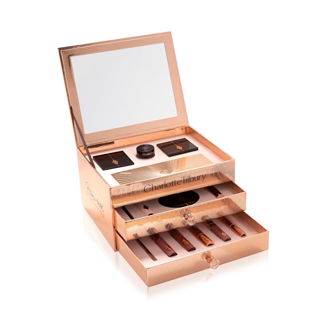 Pillow Talk Dreams Makeup Vault Box 2021 with the makeup products displayed A reflective, gold-coloured chest with two drawers containing makeup items, which include Liquid highlighter wand in rose gold, black mascara, shimmery lip gloss in nude pink, glitter-free but high-shine lip gloss in nude pink, rose gold eyeshadow pigment in a petite pot, two closed lipsticks, travel-size lip gloss in nude pink, eyeliner pencil in dark brown, lip liner pencil in nude pink, quad eyeshadow palettes, 12-pan eyeshadow palette, and two-tone blush compacts.