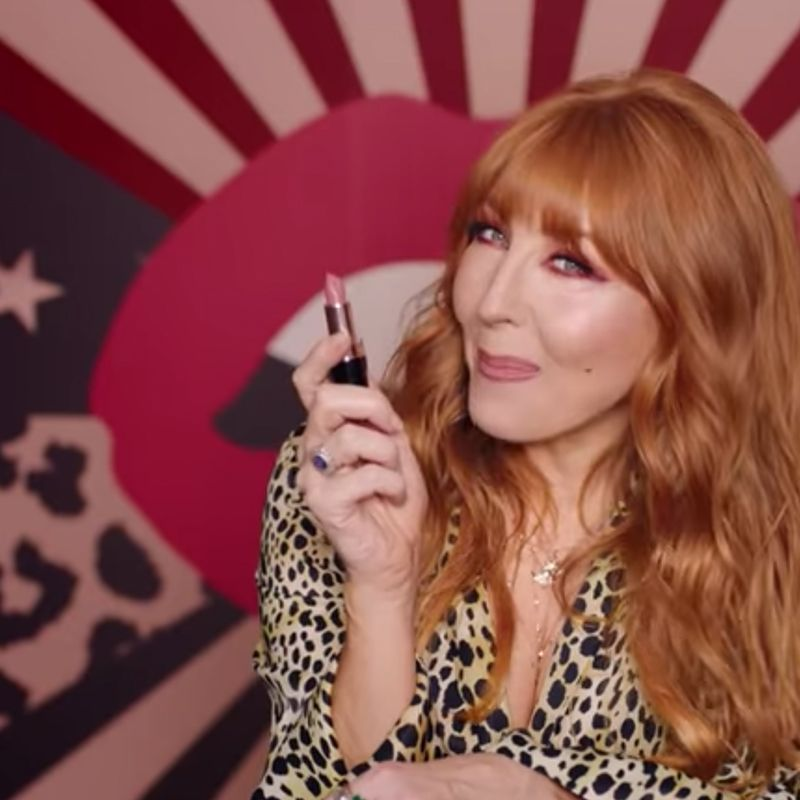 Charlotte Tilbury holding an open lipstick in a nude pink shade while wearing a leopard-print shirt with warm pink lipstick and berry-pink eye makeup. 
