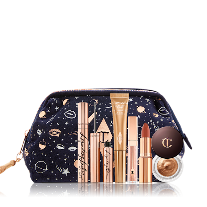 Build Your Own Makeup Kit displayed with bag A dark blue bag with a galaxy-themed print with its makeup products displayed in front of it, mascara in a gold tube, eyebrow tint in gold tube, liquid highlighter wand, nude pink lip gloss, matte lipstick, and cream eyeshadow in a glass pot.