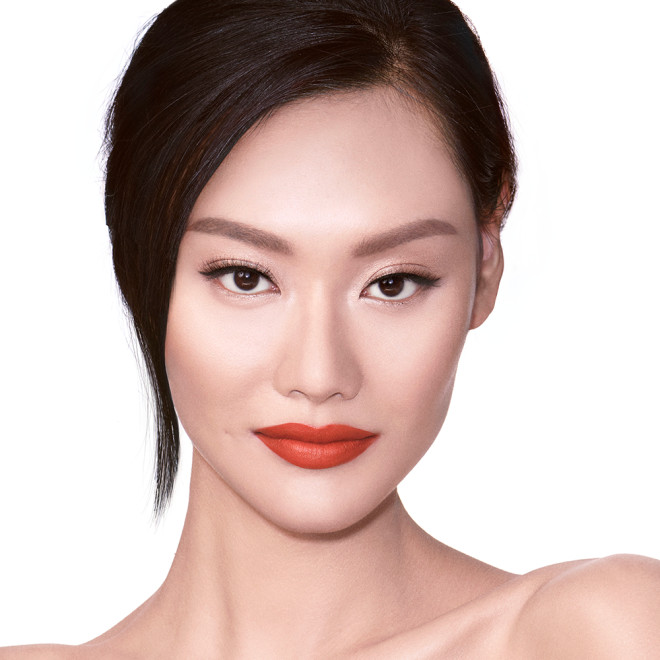 A fair-tone brunette model wearing light pink shimmery eye makeup with glowy face base and matte, orange-red lipstick. 