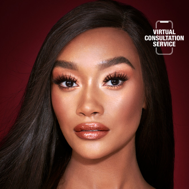 A deep-tone model with brown eyes wearing shimmery copper and gold eye makeup with black eyeliner, glowy bronzed cheeks, and pinkish-brown lipstick with gloss on top. 
