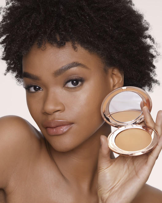 Deep-tone brunette model with flawless, matte skin wearing a nude pink lipstick and holding an open, pressed powder compact in a tan shade.