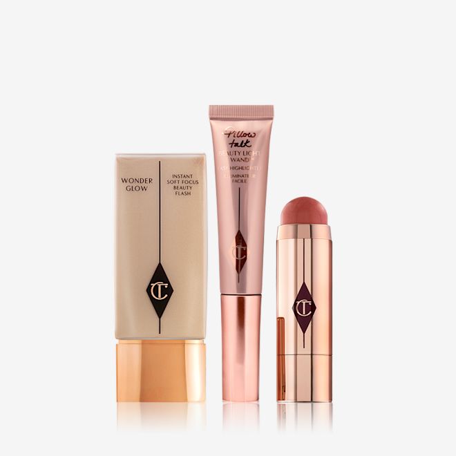 Happy Beauty Glow Kit still life A light-beige-coloured primer in a rectangular bottle with a golden-coloured lid, a highlighting wand in rose-gold packaging, and an open blush stick in a terracotta colour in a reflective, golden-coloured tube.