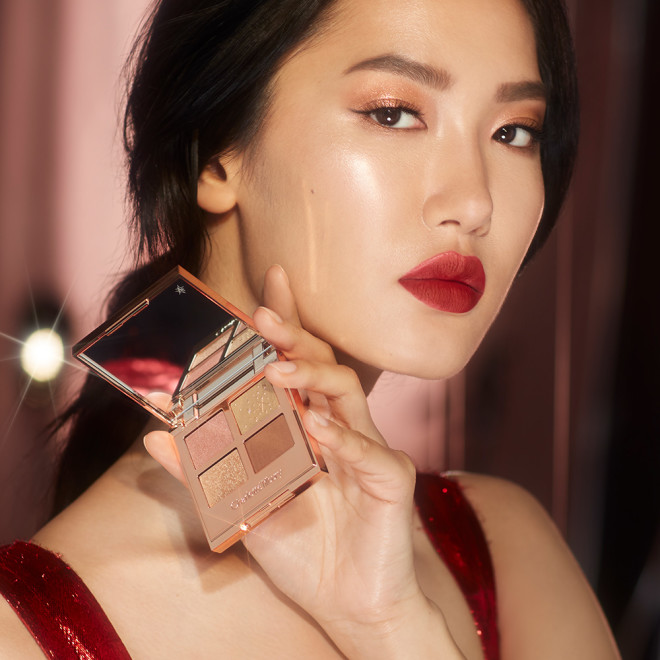 Fair-tone brunette model holding an open, quad eyeshadow palette with a mirrored lid with eyeshadows in blush pink, duochrome pink-gold, bronze brown, and pearlescent gold.