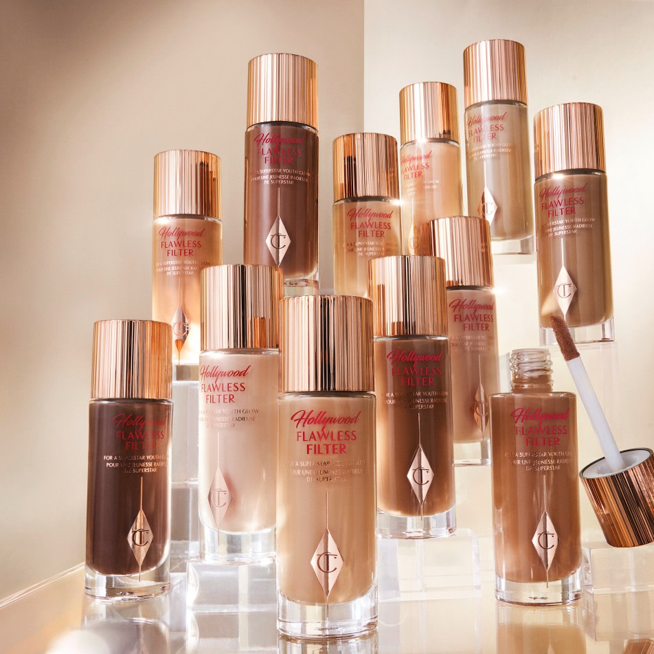 A collection of full-size glowy face primers for fair, light, medium-light, medium, medium-dark, and deep in glass bottles with gold-coloured lids and doe-foot applicators. 