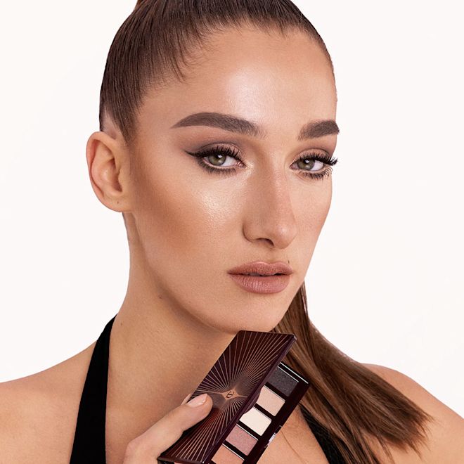 Super Nudes Easy Eye Palette on fair-tone model A fair-tone model with green eyes holding an open, mirrored-lid six-pan eyeshadow palette with matte eyeshadows in brown, peach, and beige shades while wearing a smokey brown eye look created using that palette with nude peach lipstick.