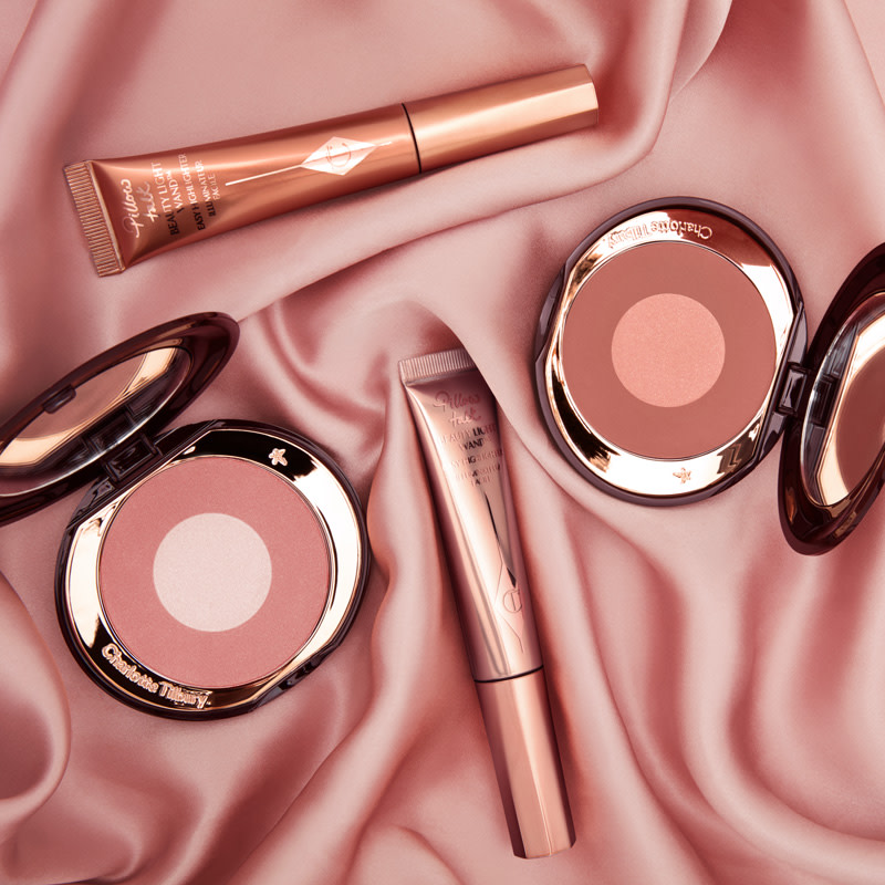 Two open, two-tone powder blushes in warm pink and brown-pink with a liquid blush and liquid highlighter wands in nude pink and bronze, placed on a nude pink silk sheet. 