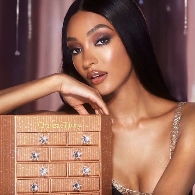 Charlotte's Instant Eye Palette Advent Calendar held by deep-tone model Banner with a golden-coloured chest of drawers filled with skincare and makeup items along with a deep-tone model carrying the chest