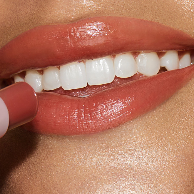 Lips close-up of a medium-tone model applying a moisturising lipstick balm in a peachy-nude shade.