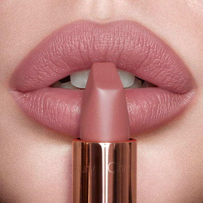Pillow Talk Lipsticks card group APAC Lips close-up of a light-toned model wearing a nude-pink matte lipstick while holding the lipstick in front of her lips.