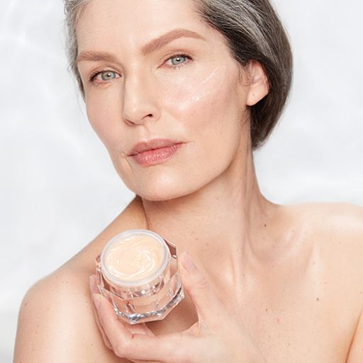 Magic Night Cream on fair-tone model with mature skin Fair-tone model with glowy, fresh, and mature skin, holding an open glass jar of a thick, peach-coloured night cream.
