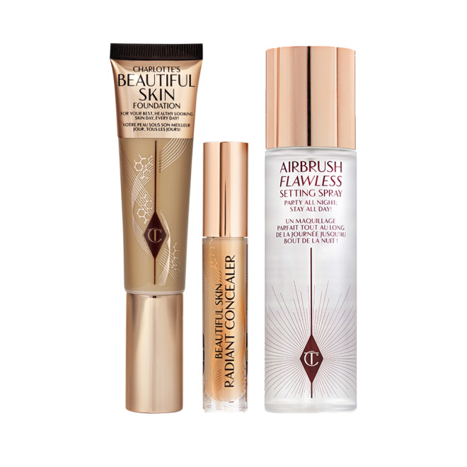Foundation in a sleek gold-coloured tube with a concealer in a glass tube and gold-coloured lid, dark brown cream bronzer compact in gold-coloured packaging, and setting spray in a large, clear bottle with a gold-coloured lid.