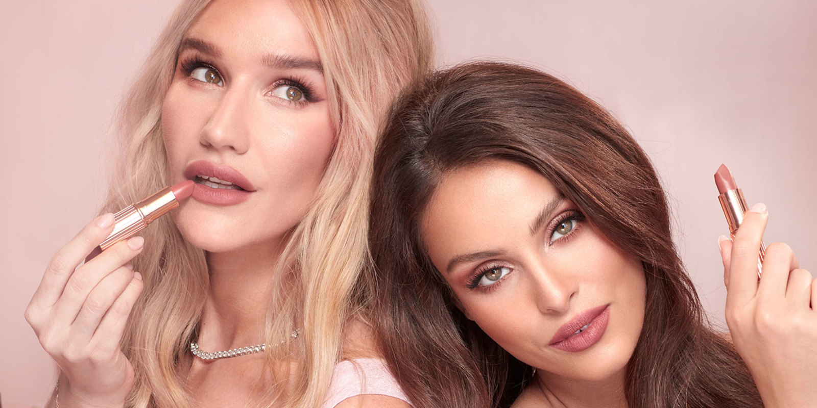 A fair-tone blonde model with a medium-tone brunette model, both wearing glowy, warm pink makeup and holding open lipsticks in warm pink shades. 