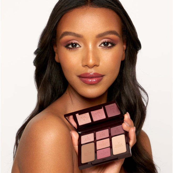 Instant Look in a Palette in Sunset Dreamscape held by deep-tone model Deep-tone brunette model with brown eyes wearing shimmery rose gold and champagne eyeshadow, glowy pinky-peach blush, soft gold highlighter, and glossy lipstick in a dark magenta colour while holding an open, face palette with a mirrored-lid with three rose gold, dusky pink, and plum-coloured eyeshadows, two blushes in soft peach and medium-pink, light brown bronzer, and soft, gold-coloured highlighter.