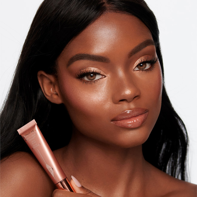 Deep-tone brunette model wearing glowy highlighter blush in a medium-pink shade while holding the tube.