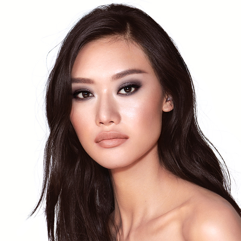 A fair-tone brunette model with brown eyes wearing smokey silvery eyeshadow with bronzed cheeks and glossy nude-peach lips. 