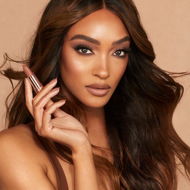 Deep-tone model with brown eyes wearing soft beige and champagne eyeshadow with a fresh, neutral nude peach lipstick with a matte finish while holding that lipstick up.