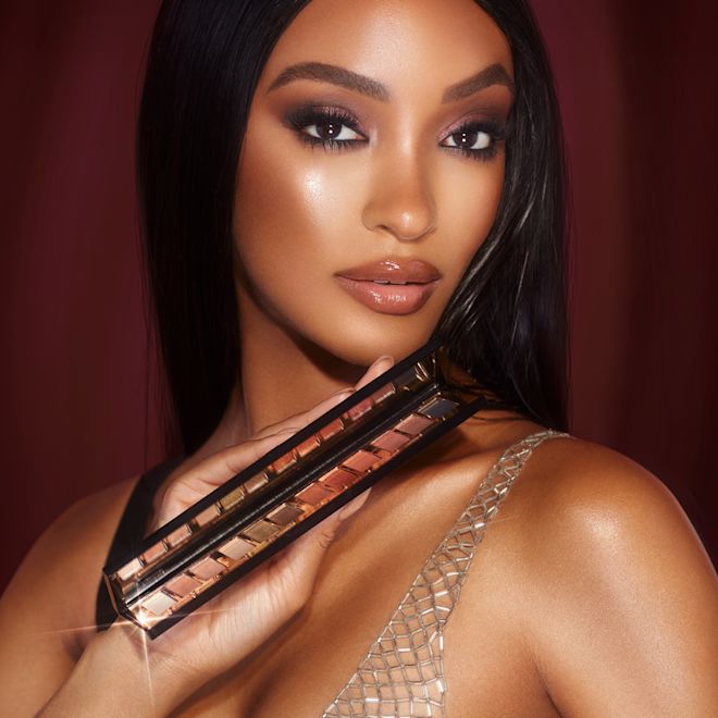 Instant Eye Palette in Smokey Eyes are Forever held by deep-tone model Deep-tone model with brown eyes wearing smokey brown eye makeup with nude pink lips and holding up an open, 12-pan eyeshadow palette with matte and shimmery shades in beige, pink, gold, green, brown, peach, and black.