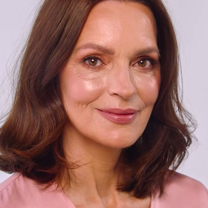 A light-tone model with mature skin wearing glowy nude pink makeup with a warm pink lipstick that brightens her face and makes it appear softer. 