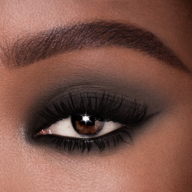 Single-eye close-up of a deep-tone model with dark brown eyes wearing jet black cream eyeshadow smudged and spread on her eyelid and the same eyeshadow used as an eyeliner for a smokey black eye look. 
