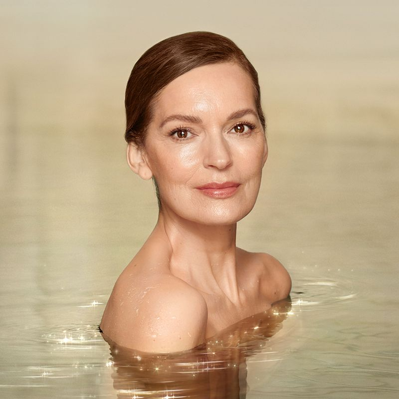 A model with mature, luminous skin, wearing nude makeup with shoulders bare, in glimmering water. 