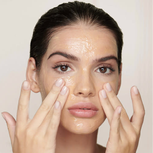 A light-tone model applying a citrusy, soft orange-coloured oil cleanser all over her face. 