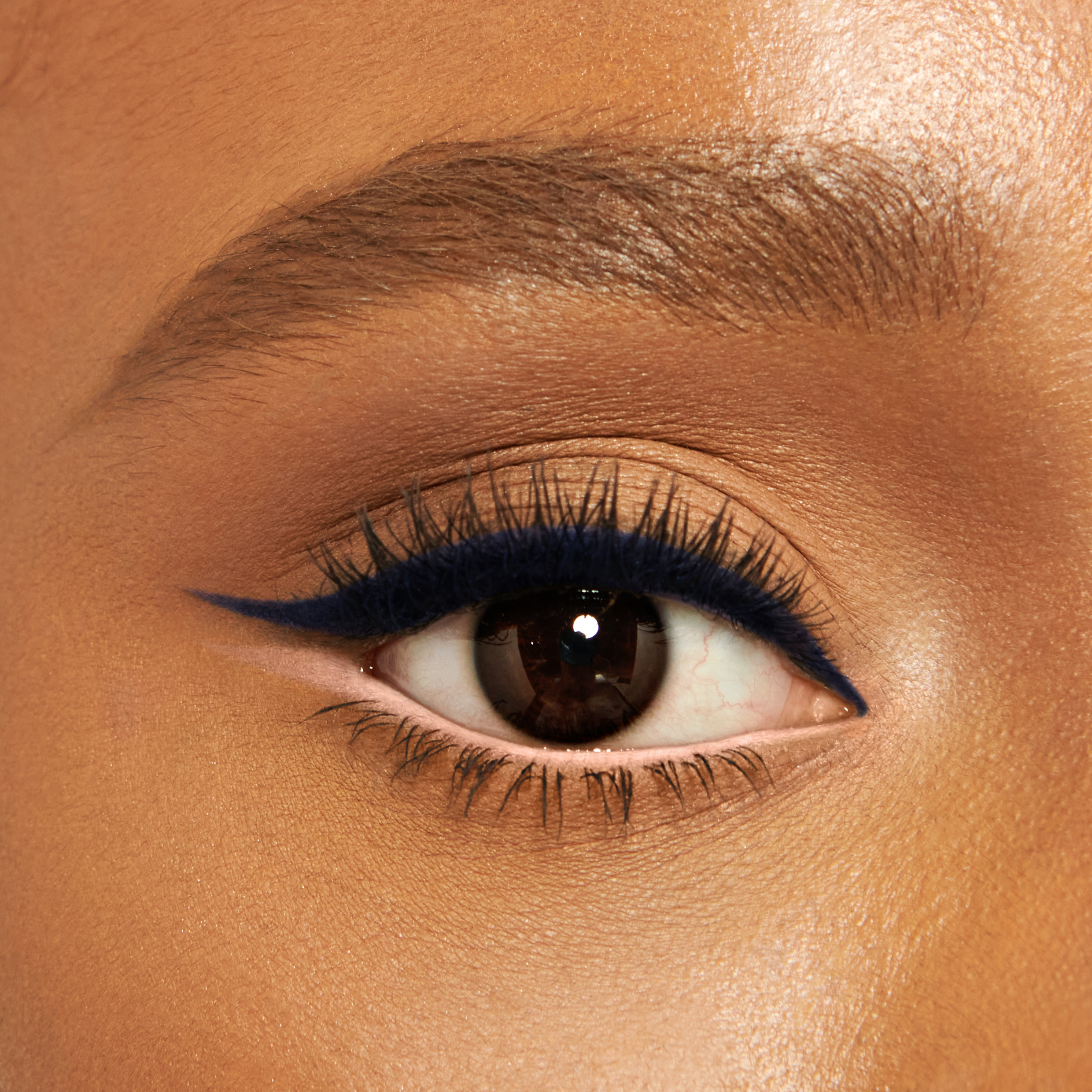 Close-up of a deep-tone model with brown eyes wearing blue-black eyeliner pencil on her eyelid and nude beige eyeliner pencil on her lower waterline. 